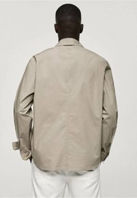 Camicia beige con un design semplice, maniche lunghe e una cucitura posteriore. Il tessuto sembra liscio, con polsini e colletto abbottonati.