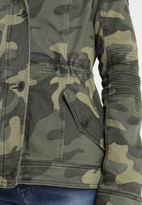 Jacket s kamuflážovým vzorem v zelených tónech, s knoflíkovým zapínáním, elastickým pasem a texturovanými detaily na rukávu. Vyrobeno z odolného materiálu.