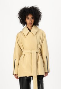 Beige bæltefrakke med en stor krave, lange �ærmer og lynlåslukninger i ærmerne. Blødt materiale med en glat tekstur, der har en afslappet pasform.