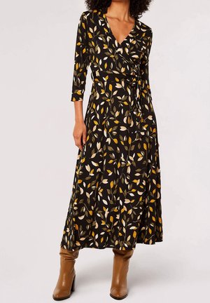 Femme portant une robe portefeuille noire avec des motifs de feuilles jaunes, blanches et vertes, associée à des bottes en cuir marron clair montant jusqu'au milieu du mollet.