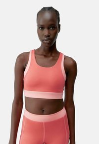 Korall rózsaszín sportmelltartó sima textúrával, széles pántokkal és testhezálló kialakítással. Zökkenőmentes élek a nyakkivágás és az alsó rész körül.