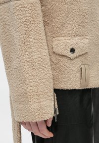 Beige geribbelde fleecejack mouw met klepzak en drukknoop, gedeeltelijk de hand en zwarte leren broek bedekkend.