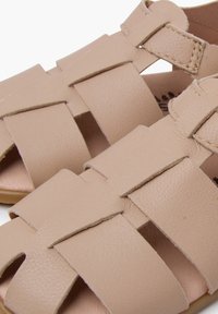 Bežid nahkkingad, millel on kootud rihmad, avatud varvaste disain ja sile tekstuur. Omab tasast tallat ja peeneid õmblusdetailidega.