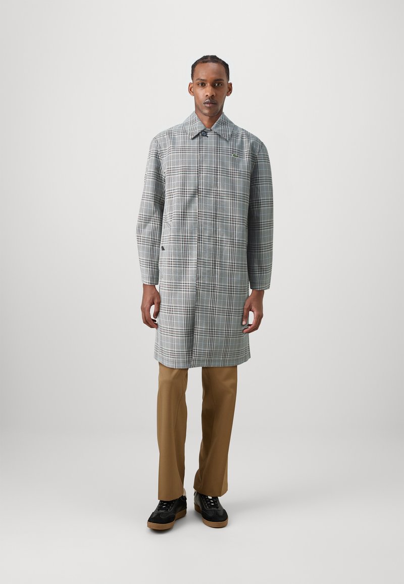 Lungo cappotto scozzese grigio e bianco con un colletto elegante, abbinato a pantaloni marroni e scarpe nere. Presenta un design minimalista e un branding sottile.