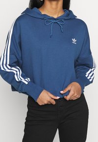 Krótka niebieska bluza z kapturem z bawełnianej mieszanki, posiada kaptur ze sznurkiem, biały logo Adidas oraz trzy białe paski na każdym rękawie.