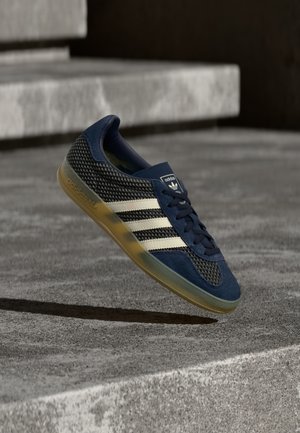 Chaussure de sport bleu marine avec dessus en mesh et daim, trois bandes blanches, et une semelle extérieure translucide couleur gomme ; modèle à lacets avec col rembourré.