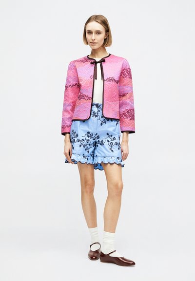 Γυναίκα που φοράει ένα ροζ υφασμάτινο σακάκι με μαύρες λεπτομέρειες, ανοιχτό μπλε κεντημένο σορτς με βολάν στο τελείωμα, λευκές κάλτσες και καφέ παπούτσια Mary Jane.