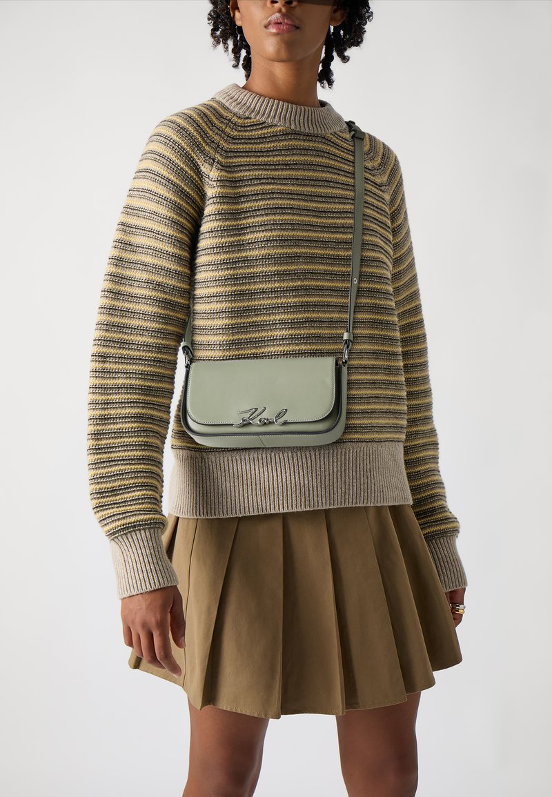 Borsa a tracolla verde in pelle con logo metallico, indossata sopra un maglione a righe e una gonna plissettata marrone. Il maglione ha un colletto a coste.