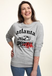 Felpa grigia con grafica nera recante la scritta "atlanta" e "SPEEDRUN" accompagnata da un'illustrazione di un'auto, con polsini e orlo a costine. Indossata con jeans blu.