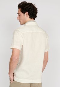 Camisa de manga corta en lino color beige claro, con un collar de patrón sutil y corte relajado. La vista trasera muestra las mangas cortas y el dobladillo recto.