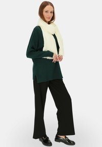 Zielony sweter z dzianiny z bocznymi rozcięciami, kremowy szalik z frędzlami, czarne spodnie z szerokimi nogawkami oraz błyszczące czarne loafersy. Białe tło.