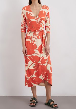 Vrouw draagt een lange mouw wikkeljurk met grote oranje bloemenprint en zwarte slippers op een effen achtergrond.