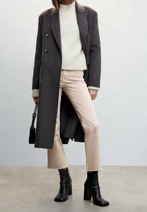Femme portant un long manteau gris foncé, un pull en maille crème, un pantalon beige clair, des bottes noires à talons, et tenant un petit sac à main noir.