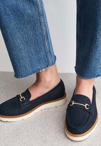 Mocassins en daim bleu marine avec un bout rond, des accents en métal doré et une semelle texturée de couleur crème. Portés avec un denim coupé.