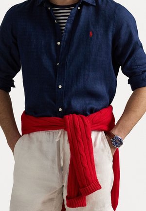 Mann trägt ein dunkelblaues Hemd über einem schwarz-weiß gestreiften Shirt, weiße Hose mit Kordelzug, roter Pullover um die Taille gebunden und Armbanduhr.