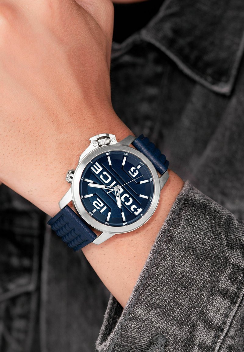 Mörkblå silikonrem klocka med silverfärgad boett, överdimensionerade vita timmarkörer och en framträdande svart urtavla med fet stil på siffrorna.