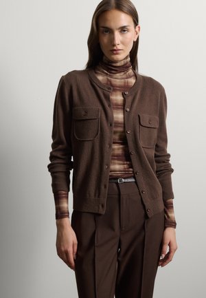 Woman wearing a brown buttoned cardigan over a striped brown turtleneck and dark brown trousers with a black belt.