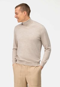 Beiger Rollkragenpullover aus glattem Strickstoff. Mit langen Ärmeln und gerippten Bündchen. Kombiniert mit hellbeigen Hosen.