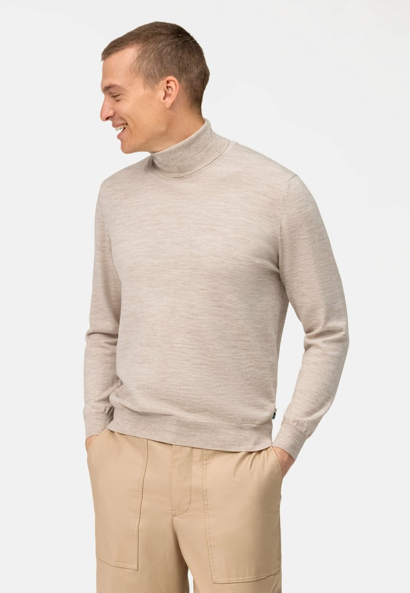 Beiger Rollkragenpullover aus glattem Strickstoff. Mit langen Ärmeln und gerippten Bündchen. Kombiniert mit hellbeigen Hosen.