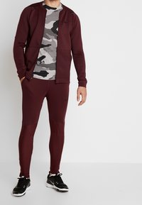 Man in een bordeauxrood Nike trainingspak met een ritsjack, een T-shirt met camouflagepatroon, en zwarte Nike sneakers, staand tegen een witte achtergrond.