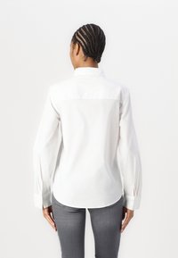 White long-sleeve button-up shirt with a relaxed fit, featuring a split detail at the cuffs and a curved hem. Smooth cotton fabric.