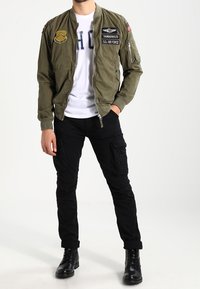 Olivgrön bomberjacka med lappar, dragkedjestängning och ribbade ärmar, bärs över en vit skjorta. Matchas med svarta cargo-byxor och svarta stövlar.