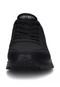 Scarpa sportiva nera con una parte superiore liscia e testurizzata, punta rotonda e lacci piatti. Presenta una suola ammortizzata e un logo sulla linguetta.