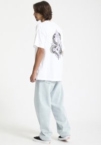 T-shirt en coton blanc avec un design graphique représentant des serpents et des épines en noir et en couleurs pastel au dos ; associé à un pantalon en denim clair.