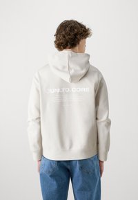 Cremefarvet hættetrøje med snoretræk i hætten, udstyret med et stort grafisk print på ryggen, parret med blå denim jeans.