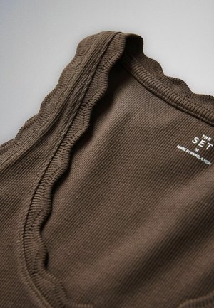 Tuvplāns brūnai ribotai auduma daļai ar viļņotu malu un baltu drukātu etiķeti ar tekstu "The Set, M, Made in Bangladesh."