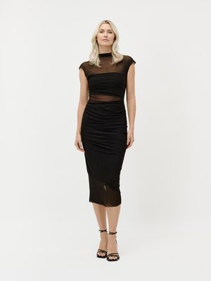 Nő áll, fekete, testhez simuló midi ruhát visel átlátszó hálós betétekkel és húzott oldalalakkal, fekete pántos, magassarkú szandálokkal párosítva.