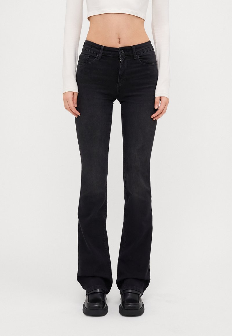 Femme portant un jean flare noir ajusté, des chaussures noires et un top court blanc à manches longues, debout devant un fond blanc uni.