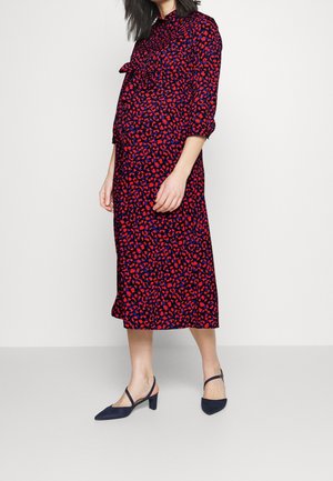 Kvinde iført en rød og blå mønstret midi-kjole med trekvartærmer og navy lavhælede slingback-sko, stående mod en ensfarvet baggrund.