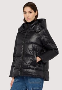 Giacca a piumino nera con finitura lucida, chiusura con zip, protezione per il mento e cappuccio ampio. Dotata di tasche laterali e design trapuntato orizzontale.