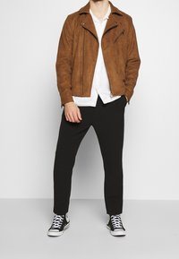 Brun ruskinds motorcykeljakke, hvid skjorte med knapper, sorte bukser og sorte højtop sneakers, står op ad en ensfarvet baggrund.