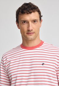 Randig t-shirt med röda och vita horisontella ränder, rund halsringning med röd kant, med en liten broderad logotyp i blått på bröstet.