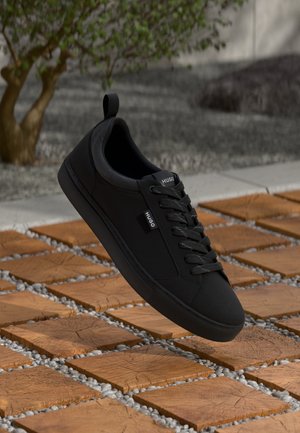 Baskets noires HUGO avec lacets sur des carreaux en bois carrés et petits cailloux blancs, plante verte en arrière-plan.
