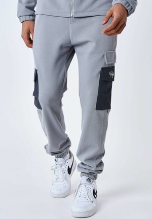 Pantalons de survêtement gris avec une finition texturée, présentant des poches cargo noires sur les côtés et des poignets élastiques. Associés à des baskets blanches.