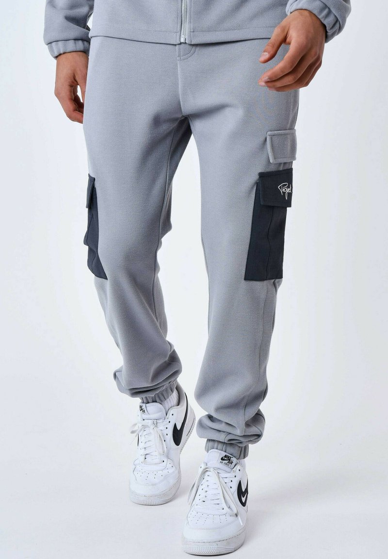 Pantaloni grigi in felpa con una finitura testurizzata, dotati di tasche cargo nere sui lati e polsini elasticizzati. Abbinati a sneaker bianche.