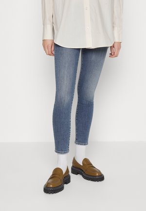 Personne portant une chemise blanche à boutons, un jean slim bleu, des chaussettes blanches et des mocassins marron à semelle épaisse, debout sur un fond uni.