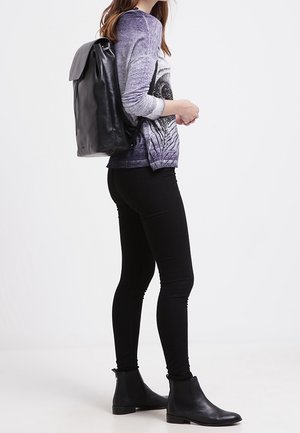 Sac à dos en cuir noir avec un rabat pliant, porté avec un pull gris à motifs et un jean skinny noir, complété par des bottines noires.