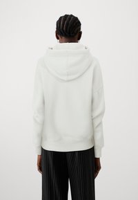 White hooded sweatshirt with a relaxed fit, ribbed cuffs, and hem. The fabric appears soft with a smooth texture, complemented by black striped trousers.