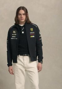 Mladic s kosom do ramena nosi crnu trkaću jaknu s logotipima, bijele hlače i sunčane naočale objesene o ovratnik svoje košulje.