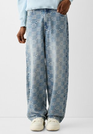 Persona che indossa jeans a gamba larga a quadretti azzurri con scarpe da ginnastica bianche e una felpa azzurra, in piedi su uno sfondo uniforme.