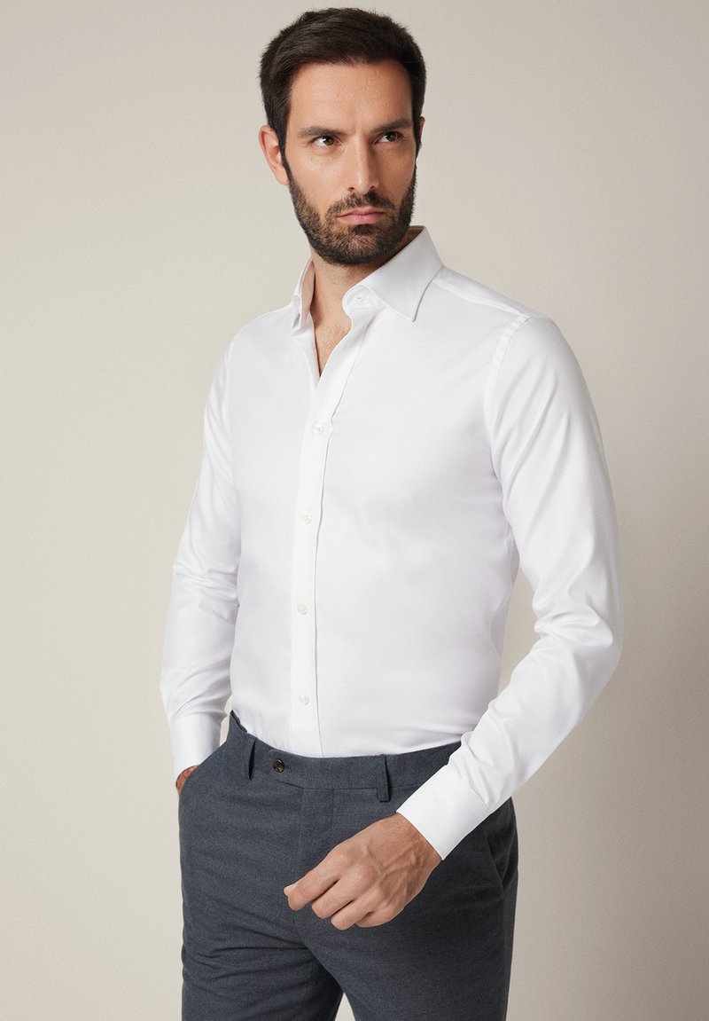 Biała, zapinana na guziki koszula na długi rękaw o gładkiej fakturze i klasycznym kołnierzyku, zestawiona z ciemnoszarymi spodniami. Krój dopasowany i dobrze skrojony.