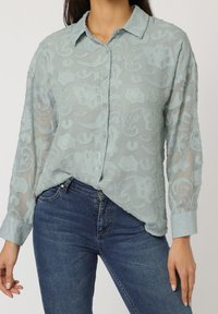 Blusa ligera azul claro con un patrón floral texturizado, cierre de botones en la parte delantera y mangas largas. Combinada con vaqueros de mezclilla azul oscuro.