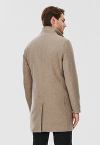 Beżowy wełniany płaszcz z wysokim kołnierzem, dopasowanym krojem i środkowym rozcięciem z tyłu. Gładka tekstura, długie rękawy i brak widocznych elementów metalowych.