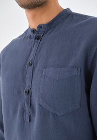 Marineblaues Hemd mit Knöpfen, das einen Stehkragen, eine Brusttasche und einen strukturierten Stoff aufweist. Sechs schwarze Knöpfe betonen das Design.