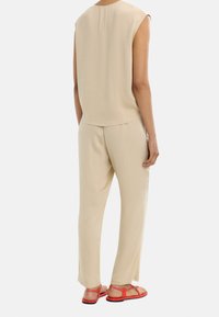 Beżowy bezrękawnik oraz dopasowane spodnie o tapered cut, wykonane z lekkiego materiału. Oferuje czerwone sandały o prostym designie paska.