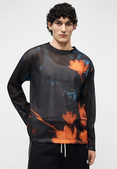 Jeune homme aux cheveux bouclés portant un haut à manches longues en maille noire orné de motifs floraux orange et bleu, associé à un pantalon noir.